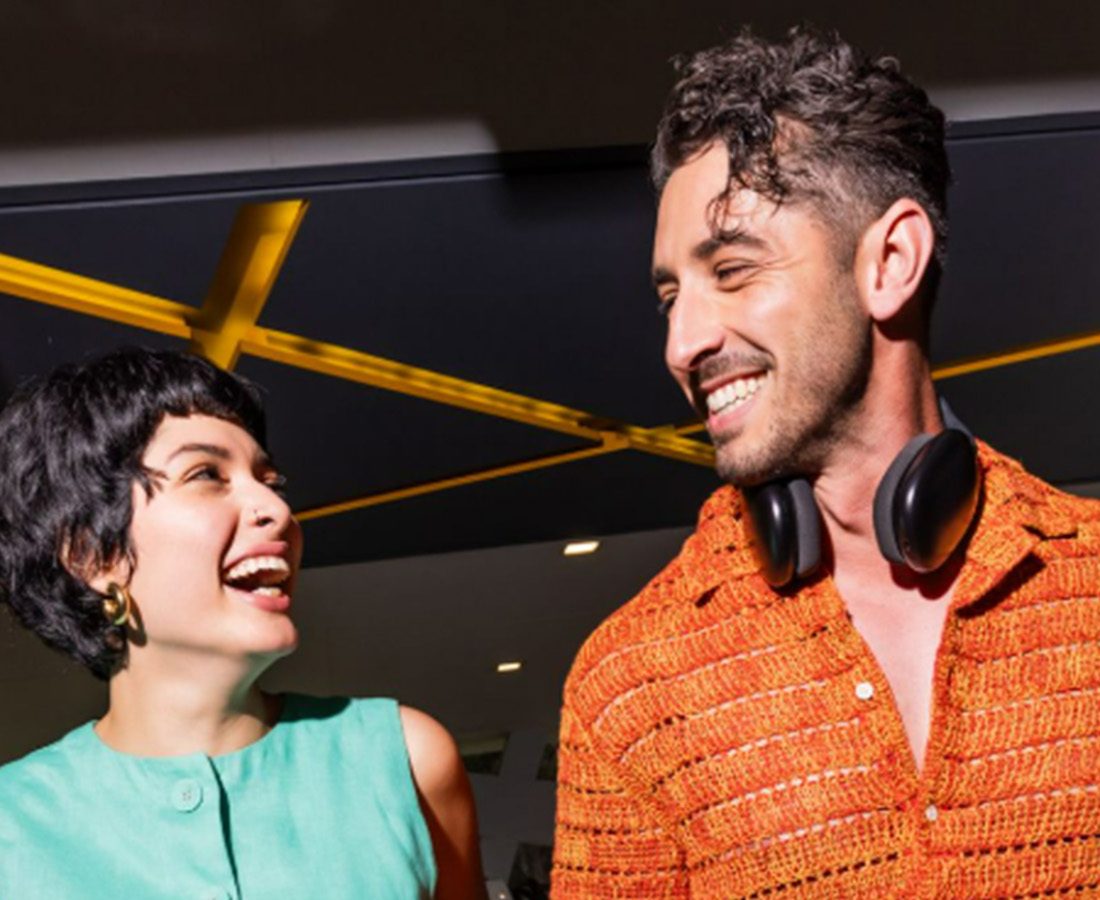 A woman in a teal sleeveless top laughs with a man in an orange shirt and headphones around his neck.
